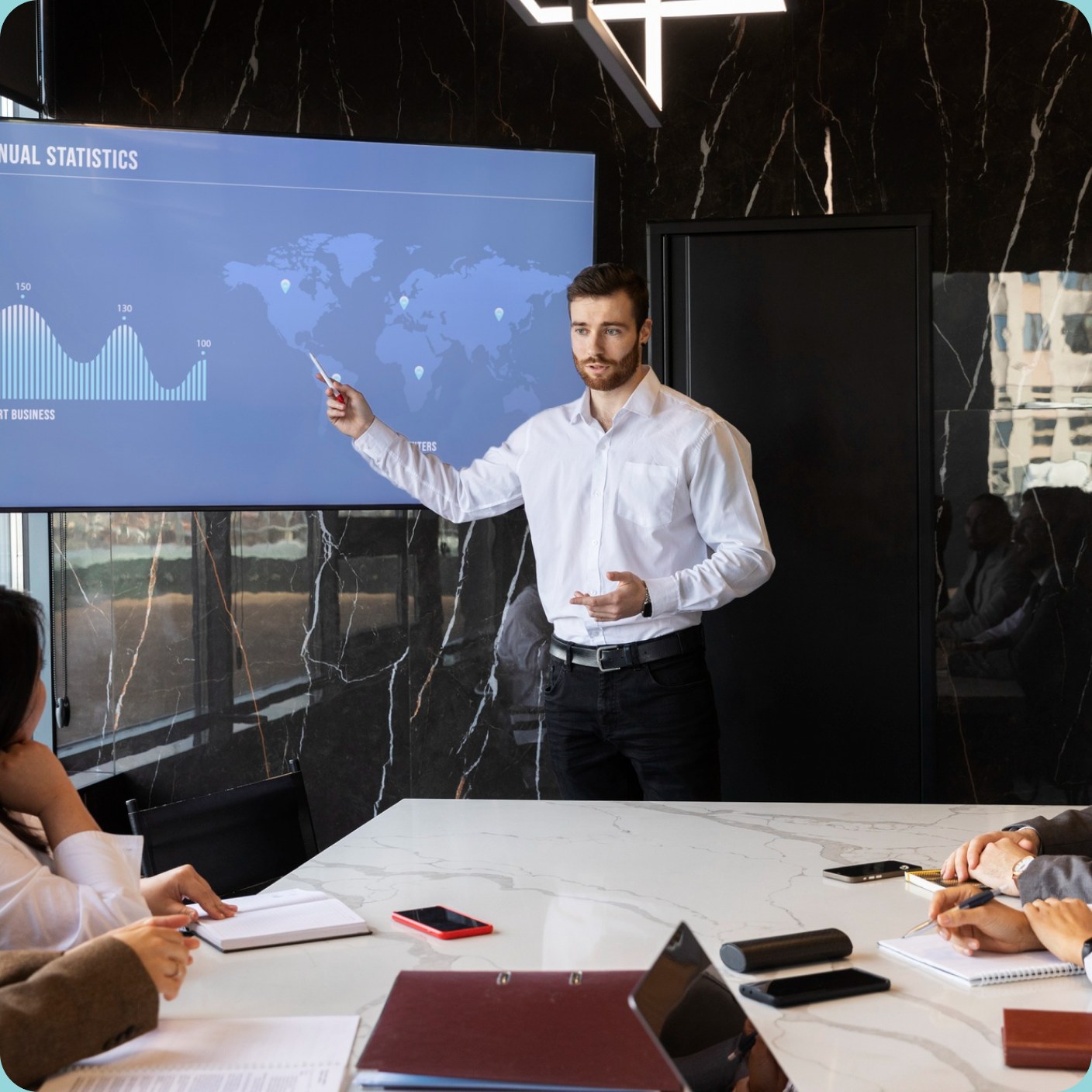 A man standing at a wall screen of annual statistics, presenting to colleagues seated around a marble meeting table.