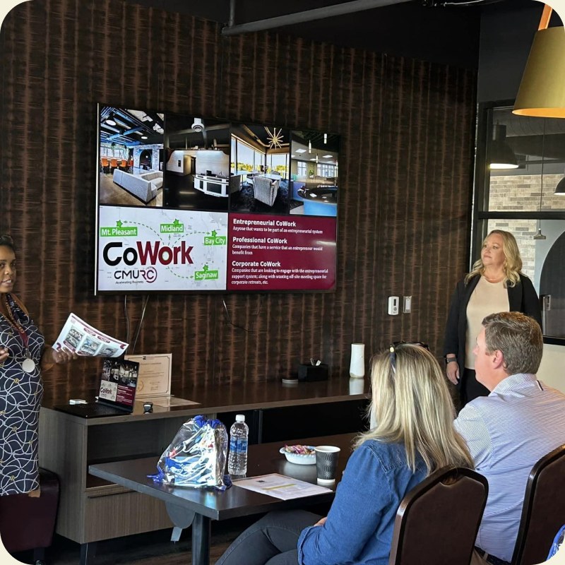 Members seated in front of a wall-mounted screen showing the CoWork display board.
