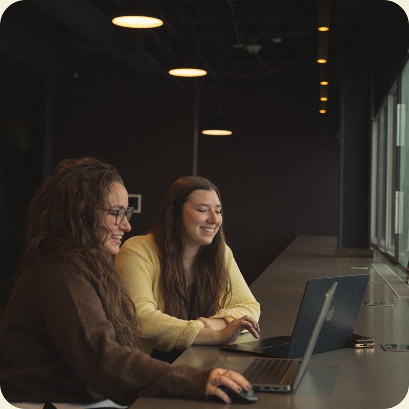 Two colleagues laughing over a laptop at a long desk in a dark-toned coworking room.