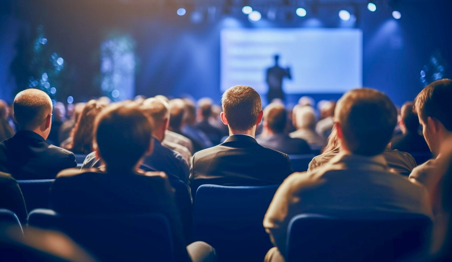 An audience in silhouette facing a brightly lit conference stage.