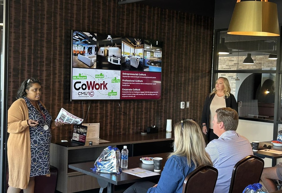 A CMURC host presenting to seated guests beside a wall screen listing the Entrepreneurial, Professional and Corporate CoWork tiers.