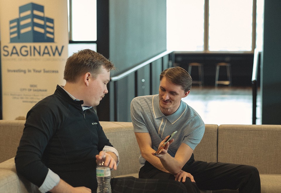 Two men talking on a sofa in front of a SAGINAW “Investing in Your Success” banner.