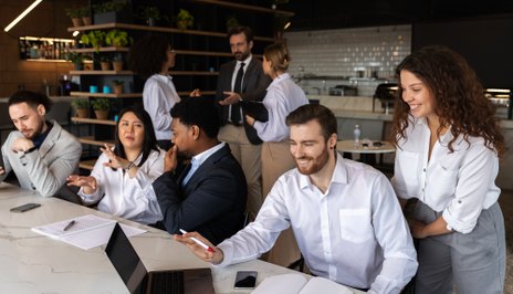 A group around a white counter in a dark, plant-filled office: a man in a white shirt gesturing at an open laptop, colleagues beside him, and a man in a suit at a whiteboard behind.