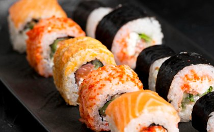 A black slate plate of sushi rolls on a dark background — salmon-topped rolls, tobiko-crusted rolls and nori-wrapped maki, shot at a low angle.