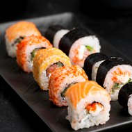 A black slate plate of sushi rolls on a dark background — salmon-topped rolls, tobiko-crusted rolls and nori-wrapped maki, shot at a low angle.