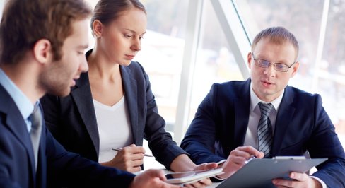 Three colleagues — two men and a woman — reviewing a tablet and printed documents at a table beside a window.