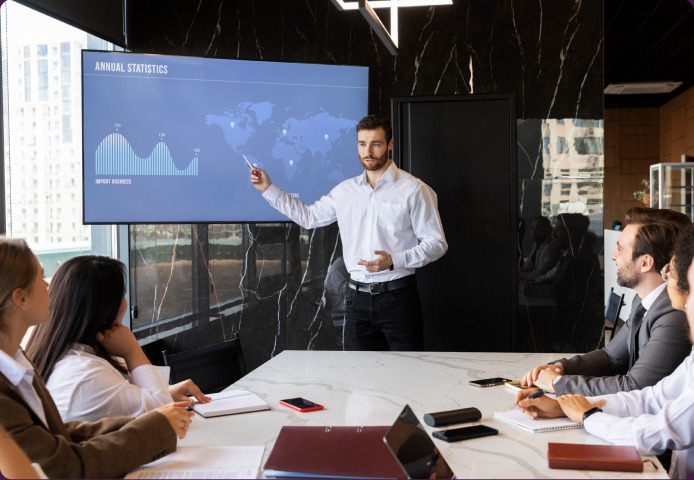 A man presenting an annual statistics dashboard on a wall screen to colleagues seated round a boardroom table.