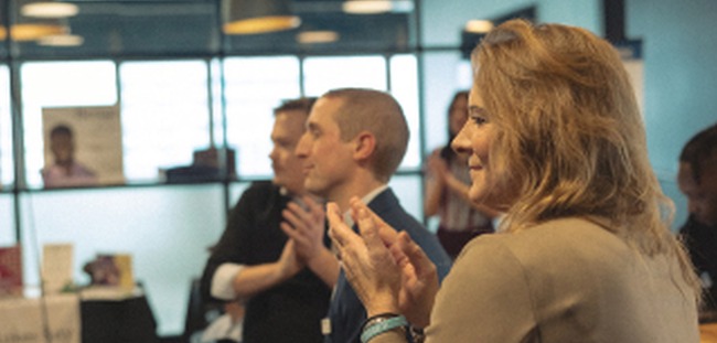 A CMURC audience applauding in a glass-walled coworking space, a woman in a tan blazer smiling in the foreground.