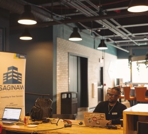 A CMURC lobby with black pendant lights, a SAGINAW pull-up banner and a person working at a desk.