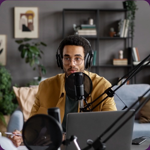 A podcast host in a mustard jacket and headphones speaking into a studio microphone.