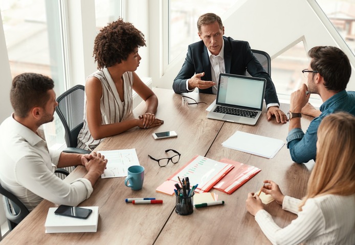 Six people meeting around a wooden table with laptops, notebooks and coffee in a daylit CMURC room.