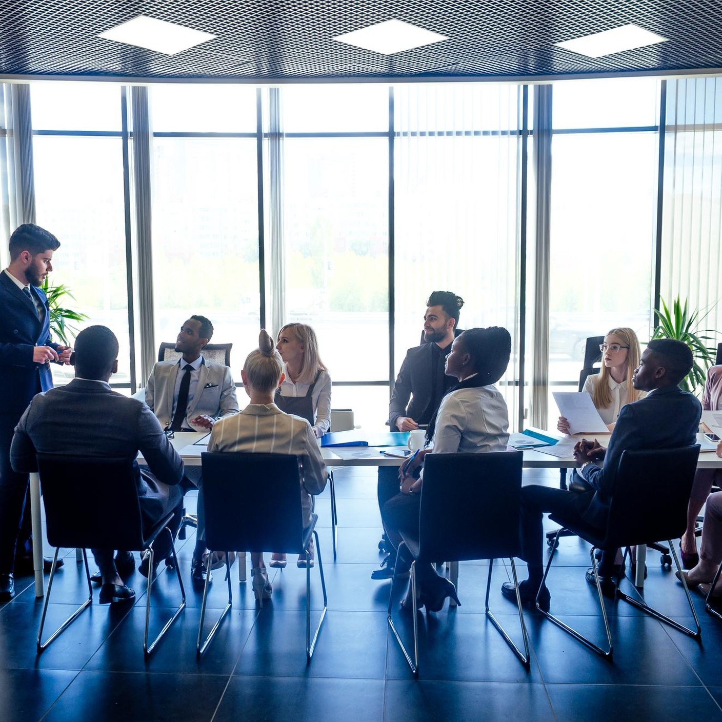 A boardroom of about eight people seated at a long table against a wall of windows, one person standing and presenting.