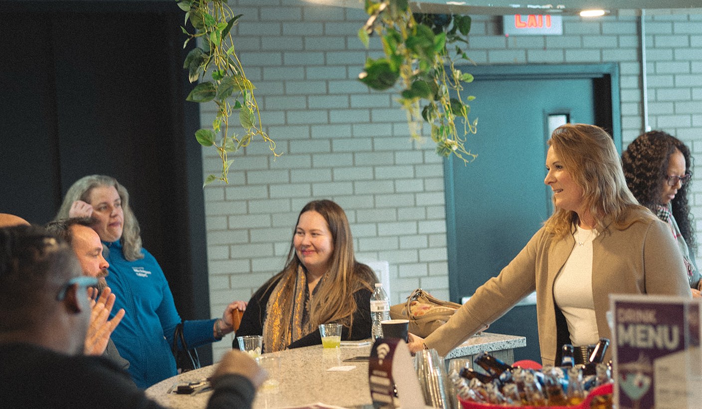 A group talking around the CMURC cafe counter, under trailing plants against a teal tiled wall.