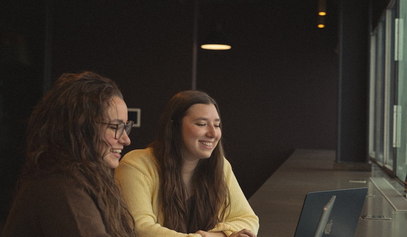 Two women sitting side by side at a desk in a dimly lit CMURC room, smiling at a monitor.