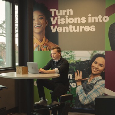 Two people at a table beside a wall graphic reading Turn Visions into Ventures.